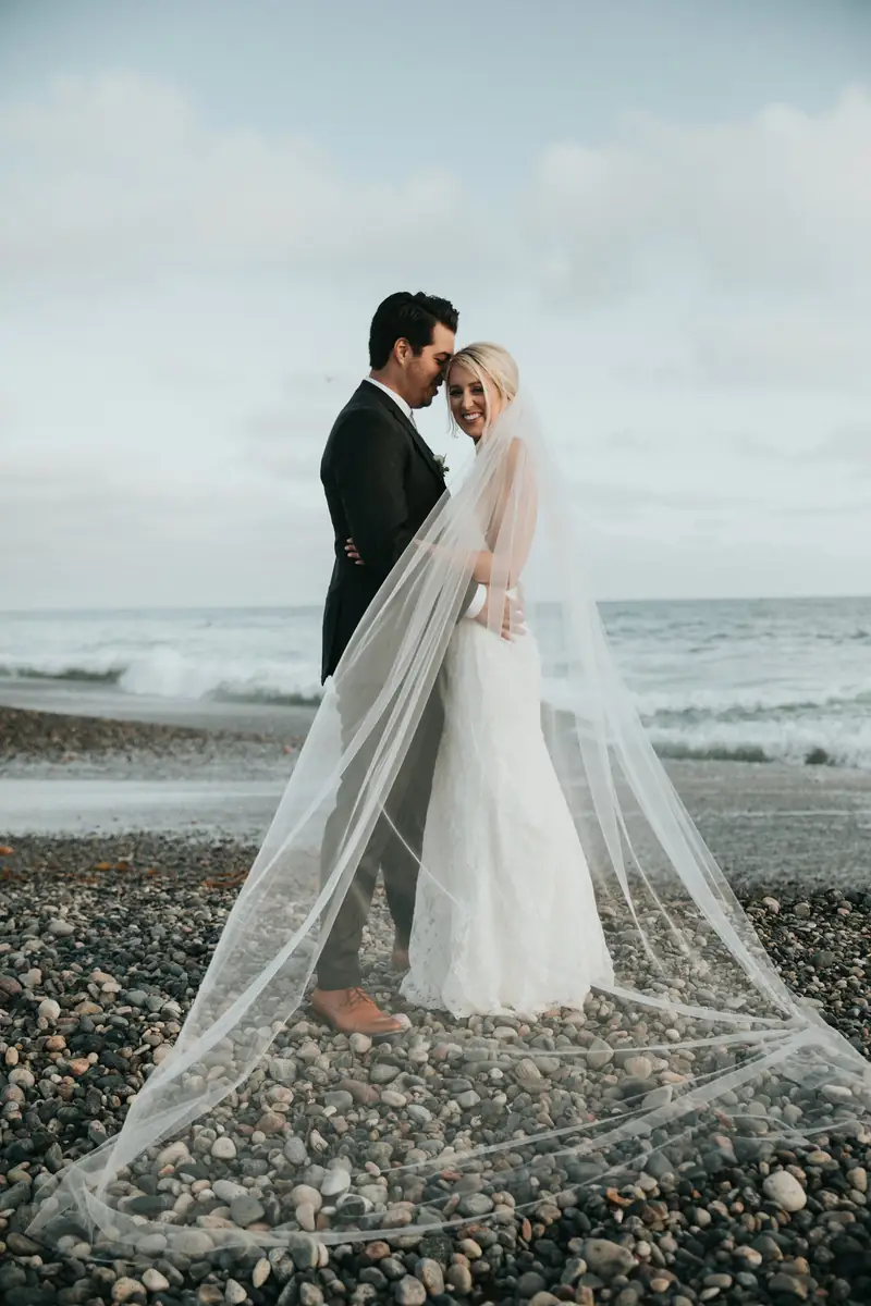 Bride and groom on a pebble beach with a long veil trailing behind